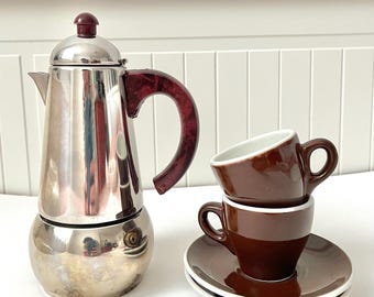 Könnte beinhalten: Eine Espressokanne aus Edelstahl mit burgunderfarbenem Griff und Deckel. Zwei braune Espressotassen und Untertassen sind daneben gestapelt. Die Tassen haben weiße Innenräume und die Untertassen einen weißen Rand.
