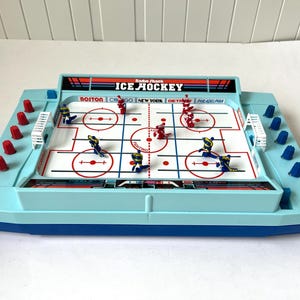 May include: A vintage Radio Shack Ice Hockey tabletop game. The game has a light blue plastic base with a white playing surface marked with red lines. Miniature hockey players in blue and red uniforms are positioned on the ice.