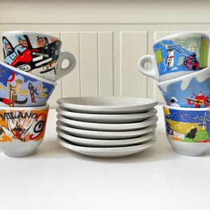 May include: A stack of six white ceramic espresso cups with colourful illustrations. The cups are stacked in two groups of three. The top cup in each stack has a handle on the right side. The bottom cup in each stack has a handle on the left side. The cups are on a white surface.
