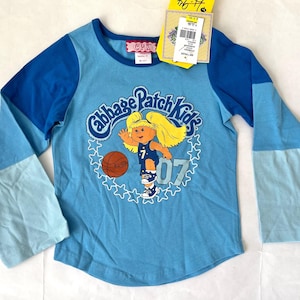 Peut inclure: Un t-shirt à manches longues bleu clair avec des manches bleues et bleu clair. Le t-shirt présente un logo Cabbage Patch Kids avec une fille jouant au basket-ball et le texte "Cabbage Patch Kids 07".