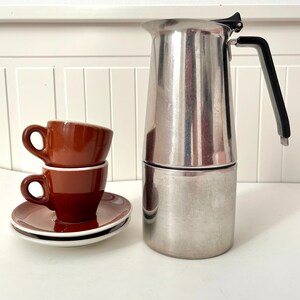 May include: A stainless steel stovetop espresso maker with a black handle and two stacked brown espresso cups with saucers. The cups and saucers are brown with a white rim.