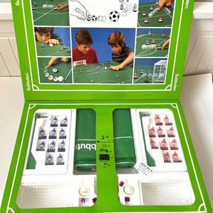 May include: A vintage Subbuteo tabletop soccer game set in a green box. The box features illustrations of gameplay and includes miniature soccer players in blue and red uniforms, a green field, goals, and accessories. The Subbuteo logo is visible.
