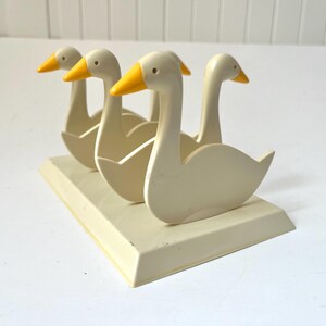 May include: A decorative goose figurine set. The set features four cream-colored geese with yellow beaks, arranged on a rectangular base. The geese are stylized with curved necks and open wings, creating a charming, vintage aesthetic.