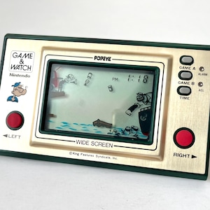 Könnte beinhalten: Ein Vintage Nintendo Game & Watch Handheld-Elektronikspiel mit der Figur Popeye. Das Spiel hat einen gold-grünen Rahmen, einen Bildschirm mit einer Popeye-Szene und rote Steuertasten. Die Uhrzeit wird als 13:18 Uhr angezeigt.