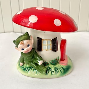 Könnte beinhalten: Ein skurriles Keramik-Pilzhaus mit rotem Hut und weißen Punkten. Eine grün gekleidete Elfenfigur liegt auf der grasbewachsenen Basis. Das Haus hat ein Fenster mit Fensterläden und einen rosa Briefkasten, was ein Märchenmotiv hervorruft.