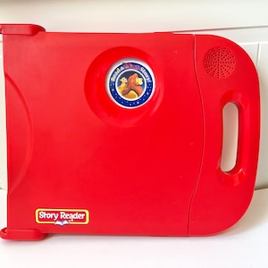 Peut inclure: Un jouet Story Reader rouge vif avec une poignée et un haut-parleur. Il comporte un emblème circulaire avec un lion et les mots "Read a Story". Les mots "Story Reader" sont imprimés en bas.