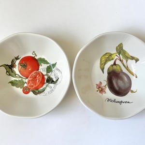 Może przedstawiać: Dwie białe ceramiczne miski z ilustracjami owoców. Jedna miska przedstawia pomidora z zielonymi liśćmi, a druga bakłażana ze słowem "Melanzana". Miski są okrągłe z lekko falistą krawędzią.