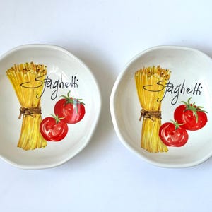 Pode incluir: Duas tigelas de cerâmica branco-sujo com um design de espaguete e tomate. Cada tigela apresenta um feixe de espaguete amarelo amarrado com uma corda marrom, dois tomates vermelhos e a palavra "Spaghetti" em escrita preta.