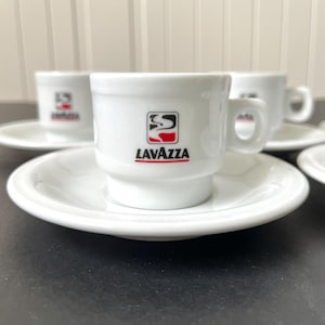 Pode incluir: Um conjunto de chávenas de café expresso e pires de cerâmica branca. Cada chávena apresenta o logótipo "LAVAZZA" em preto, vermelho e branco. As chávenas estão empilhadas nos seus pires, com mais algumas no fundo. O conjunto está numa superfície escura.