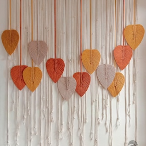 May include: A macrame wall hanging with a white background and a variety of leaves in shades of orange, yellow, and beige. The leaves are made of yarn and are hanging from a wooden dowel.