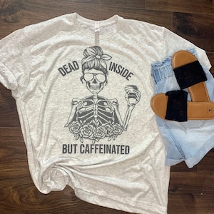May include: A light gray t-shirt with a graphic of a skeleton holding a cup of coffee. The text on the shirt reads "Dead Inside But Caffeinated".