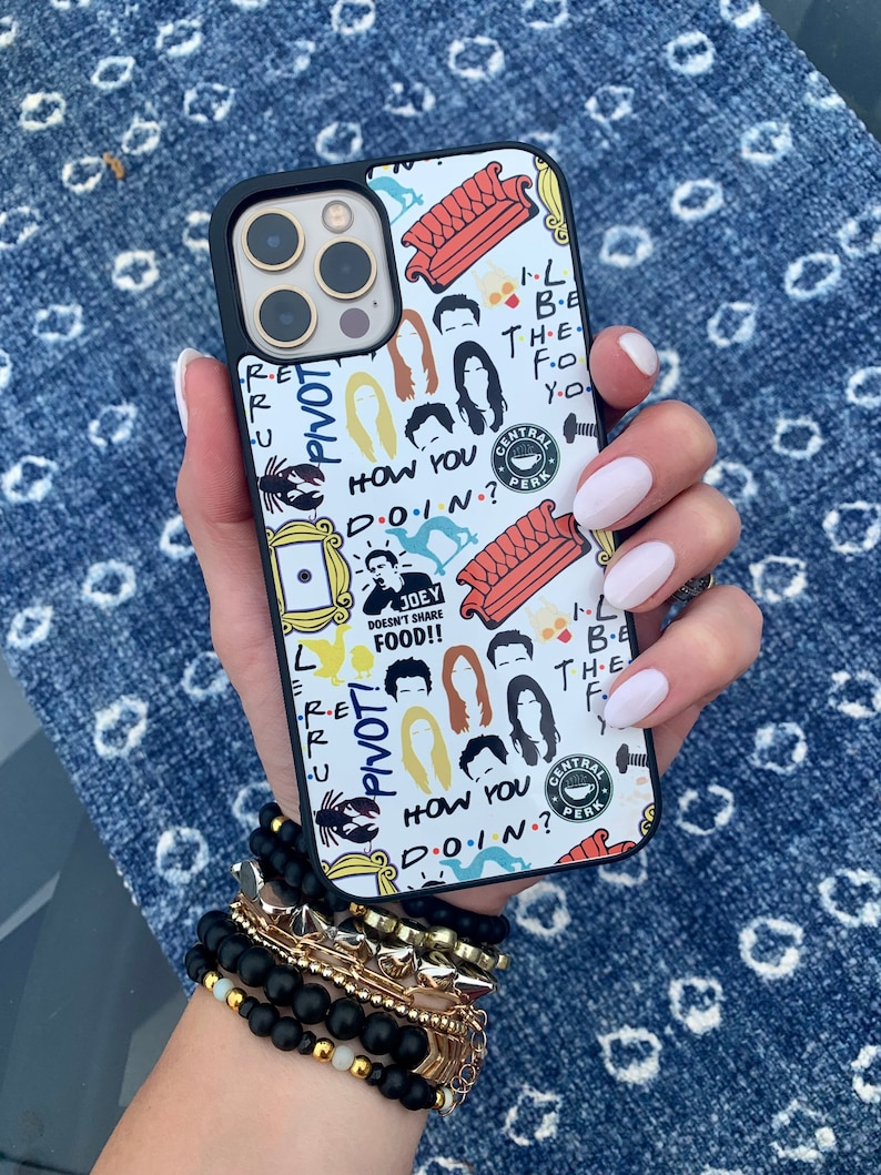 May include: A white iPhone case with a black border featuring a Friends theme. The case is covered in colorful graphics, including the iconic orange couch, the Central Perk logo, and the characters' faces. The text "Pivot!" and "How you doin?" are also featured on the case.