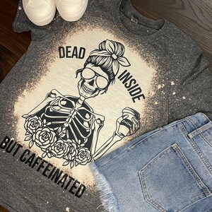 May include: A gray t-shirt with a bleached design featuring a skeleton holding a coffee cup. The text on the shirt reads "Dead Inside But Caffeinated".