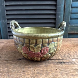 May include: A ceramic basket-shaped planter with two handles. The planter is painted in shades of green and brown, with a woven basket design. Red roses and green leaves decorate the front of the planter. The planter sits on a wooden surface.