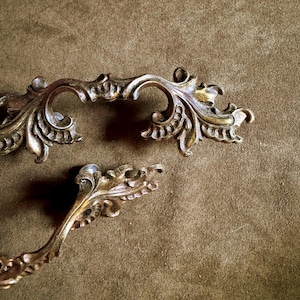 May include: Two ornate drawer pulls crafted from a metallic material with a weathered gold finish. Each handle features intricate, raised scrollwork and leaf-like detailing along the edges, with a curved shape and visible mounting points.