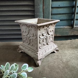 May include: A square-shaped planter with a light beige, matte finish. The exterior features raised, swirling scrollwork patterns on all four sides. The base is supported by four ornate, carved feet, and the rim has a flared, decorative edge.