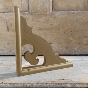 May include: A wooden shelf bracket with a decorative, ornate design. The bracket is painted a light gold colour and has a simple, rectangular base.