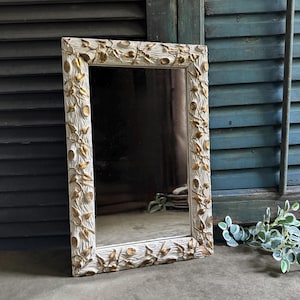 May include: A rectangular mirror featuring a textured white frame adorned with raised gold-colored leaf and vine motifs. The frame has a distressed finish, and the mirror glass reflects the surrounding environment with a clear, reflective surface.