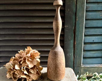 May include: A tall, light brown wooden pestle with a rounded top, a slender neck, and a wide, cylindrical base. The wood surface shows a weathered, textured grain throughout. Beside the pestle sits a cluster of dried, light brown hydrangea flowers.