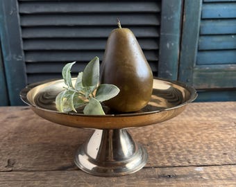 May include: A brass pedestal dish holds a faux pear and sprig of greenery. The dish has a scalloped edge and a round base. The pear is brown and the greenery is light green. The background is a weathered wooden surface.