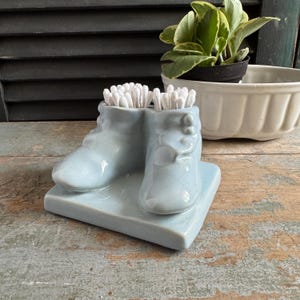 May include: A pale blue ceramic boot-shaped container filled with cotton buds. The container sits on a matching square base. A small plant in a black pot and a white bowl are in the background.