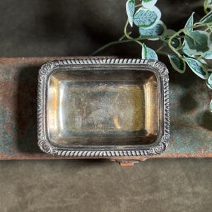 May include: A rectangular silver-toned metal tray featuring a decorative rope-style border with ornate scrollwork details at each corner. The interior surface of the tray shows a smooth, reflective finish with visible signs of wear and patina.