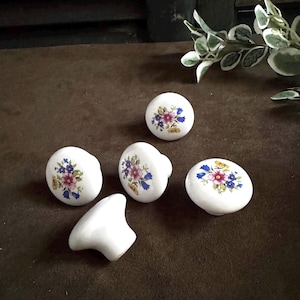 May include: A set of four round white ceramic cabinet knobs, each featuring a colorful floral pattern with pink, blue, and yellow blossoms. Three knobs are positioned with the floral design facing upward, while one knob is turned on its side.