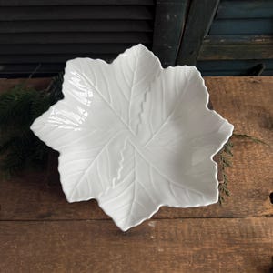 May include: A white ceramic leaf-shaped serving dish. The dish has detailed veining and a scalloped edge. The dish is placed on a wooden surface with a dark green background.