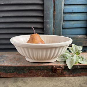 May include: A cream-colored, fluted ceramic bowl holds a brown pear. The bowl sits on a weathered, metal surface, with a sprig of green leaves to the right. The background features blue shutters and a dark, slatted blind.