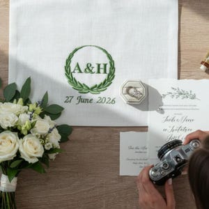 May include: A white fabric cloth featuring green embroidery of a laurel wreath surrounding the letters A&H and the date 27 June 2026. Beside it sits a ring in a beige octagonal box, a white wedding invitation, and a bouquet of white roses.