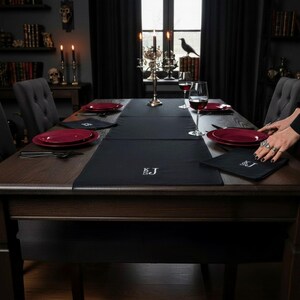 Peut inclure: Une table de salle à manger sombrement décorée. Elle est ornée de sets de table et de serviettes noirs, d'assiettes bordeaux et de verres à vin. Un chemin de table noir avec des initiales brodées est disposé au centre. La pièce est décorée de bougies et d'objets sombres.