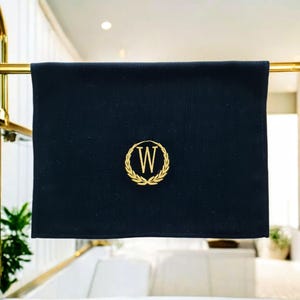 Könnte beinhalten: Ein schwarzes Handtuch mit einem goldgestickten Monogramm des Buchstabens "W", umgeben von einem Lorbeerkranz. Das Handtuch hängt an einer goldenen Stange, mit einem Badezimmer im Hintergrund.