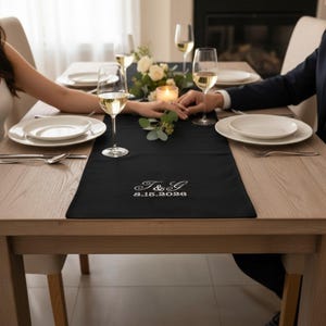 Peut inclure: Un chemin de table rectangulaire noir avec une broderie blanche indiquant T & G et la date 8.15.2026. Il est disposé sur une table à manger en bois clair avec des assiettes blanches, des verres à vin transparents et un centre de table floral.