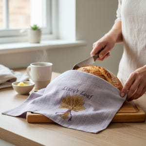 May include: A light purple linen bread cover featuring an embroidered golden wheat sheaf and the text Lizzy's Loaf in dark purple thread. The fabric is draped over a round loaf of crusty bread resting on a wooden cutting board.