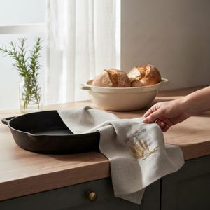 May include: A light beige linen kitchen towel featuring an embroidered golden wheat stalk design and the text "My Home Farm" draped over a black cast iron skillet. The towel is positioned near a ceramic bowl containing two round loaves of bread.