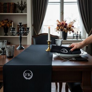 Peut inclure: Chemin de table noir avec initiales brodées blanches et motif de couronne de laurier. Sur une table en bois foncé, avec de la verrerie, un chandelier et une bougie allumée. Une main pose une serviette pliée sur une assiette.