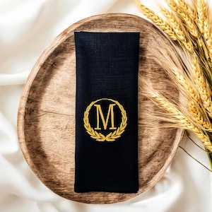 Puede incluir: Una servilleta de lino negro con la letra "M" dorada bordada dentro de una corona de laurel. La servilleta está colocada sobre un plato de madera, con espigas de trigo en el fondo. La servilleta está doblada y lista para usar.