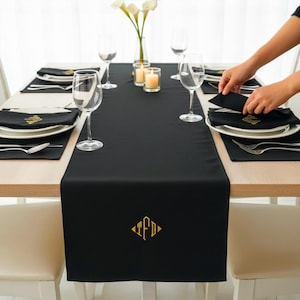 Peut inclure: Une table de salle à manger formelle avec un chemin de table noir et des serviettes assorties. La table est dressée avec des assiettes blanches, des couverts et des verres à vin. Le chemin de table présente un motif monogramme doré. Des bougies et des fleurs ajoutent à l'ambiance élégante.
