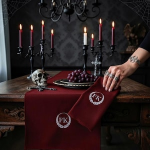 Peut inclure: Une nappe et une serviette rouge foncé brodées des initiales "FK" en blanc. La table est ornée d'un crâne, de raisins et de bougies allumées dans des candélabres noirs, créant une ambiance gothique.