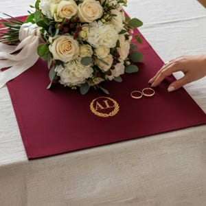 Może przedstawiać: Ciemnobordowa prostokątna mata z tkaniny ze złotym haftowanym emblematem wieńca laurowego z literami AL pośrodku. Bukiet białych i kremowych róż z zielonymi liśćmi oraz białą wstążką leży na macie obok dwóch złotych pierścionków.