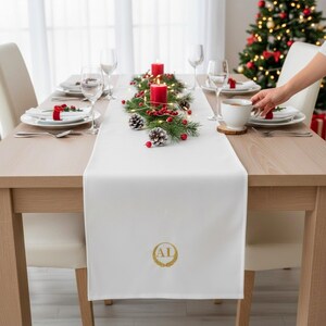 Peut inclure: Un chemin de table blanc orné d'un monogramme circulaire brodé en or avec les lettres AL. Le chemin est disposé sur une table à manger en bois avec des assiettes blanches, des verres transparents, une tasse blanche et un centre de table.