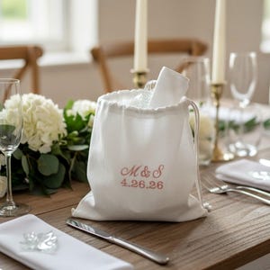 Puede incluir: Una bolsa blanca con cordón con las iniciales bordadas "M & S" y la fecha "4.26.26" en rosa. La bolsa contiene un vaso y una servilleta blanca doblada. La bolsa está sobre una mesa de madera, parte de una mesa de boda.