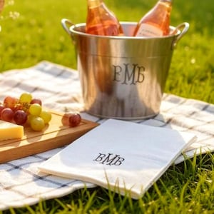 Puede incluir: Una cubeta de hielo de acero inoxidable con dos botellas de vino rosado, uvas, queso y una servilleta blanca con las iniciales "BMB" sobre una manta de picnic. La cubeta de hielo tiene las iniciales "BMB".