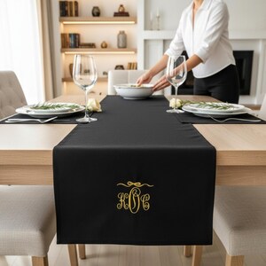 Peut inclure: Chemin de table noir avec monogramme doré, posé sur une table en bois clair. Le chemin de table est brodé des lettres KBC dans une police décorative. La table est dressée avec des assiettes, des verres et un bol blanc.