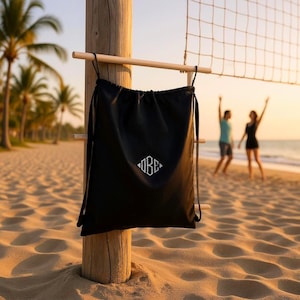 Può includere: Borsa nera con coulisse e design geometrico bianco, appesa a un palo di legno. La borsa è su una spiaggia sabbiosa con una rete da pallavolo e due persone sullo sfondo. Sono visibili anche palme e l'oceano.