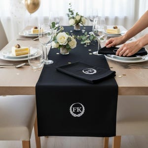 Peut inclure: Une table de salle à manger formelle avec un chemin de table noir et des serviettes assorties, chacune brodée des initiales "FK" dans une couronne de laurier. La table est dressée avec des assiettes blanches, des couverts et des verres à vin. Un arrangement floral de roses blanches et de verdure est au centre.