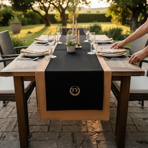 Peut inclure: Une table en bois dressée pour un repas en plein air. Elle est recouverte d'un chemin de table noir et beige avec les initiales "TY" dans une couronne dorée. Des assiettes, des couverts et des verres à vin sont disposés. Des bougies et une plante succulente sont au centre.