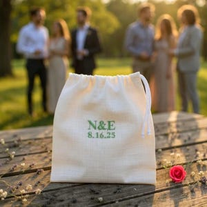 Puede incluir: Una bolsa crema con cordón, bordada con las iniciales verdes "N&E" y la fecha "8.16.25". La bolsa está sobre una superficie de madera, con una rosa roja y pequeñas flores blancas. Hay personas borrosas en el fondo.