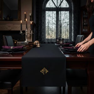 Peut inclure: Chemin de table noir avec un motif monogramme doré, posé sur une table en bois sombre. La table est dressée avec des assiettes noires, des serviettes bordeaux et de la verrerie ornée. Un chandelier argenté avec des bougies noires et un crâne complètent le thème gothique.