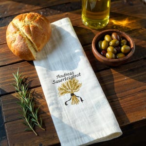 Puede incluir: Naturaleza muerta rústica con un pan redondo, una ramita de romero y un cuenco de madera con aceitunas. También hay una toalla de lino blanca con el texto "Andreas Sauerteighrot" y un diseño de trigo. Una botella de aceite de oliva está en el fondo.
